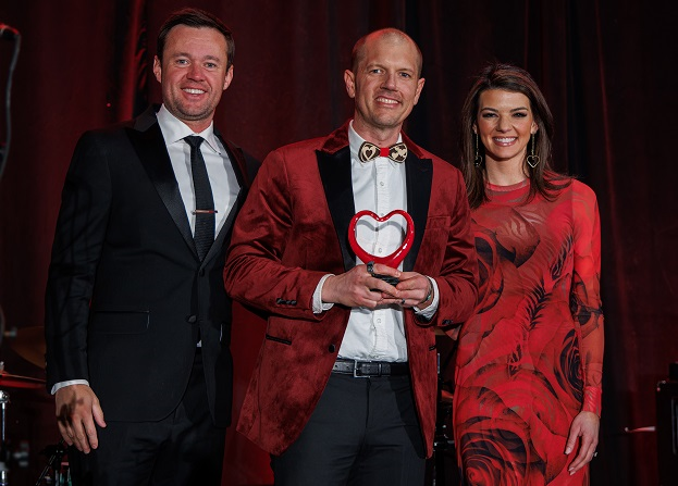 Two men and a woman on stage holding heart shaped award